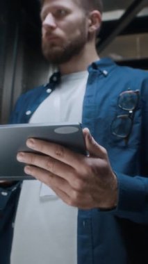 Dijital tablete bakan ve modern veri merkezindeki raf sunucu dolaplarının çalışmalarını analiz eden Kafkas erkek bilişim mühendisi dikey düşük açılı görüntüsü.