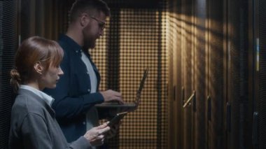 Genç beyaz kadın bilişim uzmanı, erkek arkadaşına tabletle ilgili bilgi veriyor. Dizüstü bilgisayarını kullanarak rafları kontrol ediyor. Modern veri merkezinde birlikte çalışıyorlar.