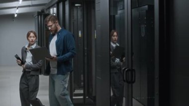Orta uzunlukta beyaz kadın bilişim uzmanı elinde tabletle sunucu odasına girerken erkek meslektaşının dizüstü bilgisayarını tutarak veri merkezindeki işi tartışması.