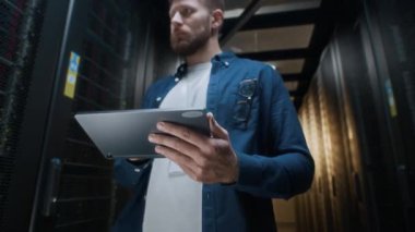 Düşük açı, dijital tablete bakıp modern veri merkezindeki raf sunucu dolaplarının çalışmalarını analiz eden, günlük giysiler içindeki beyaz erkek bilişim mühendisinin düşük açılı görüntüsü.