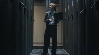 Modern veri merkezindeki raf sunucu dolaplarının çalışmalarını analiz eden günlük giysi içinde genç beyaz kadın bilişim yöneticisiyle dolu bir fotoğraf.