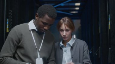 Genç siyahi erkek bilişim uzmanı dizüstü bilgisayar kullanıyor ve veri merkezindeki sunucu odasında genç beyaz kadın meslektaşla birlikte çalışıyor.