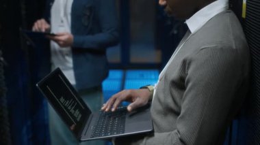 Yan görüntüde, beyaz erkek meslektaşı arka planda tablet üzerinde çalışırken genç siyahi erkek bilişim uzmanının dizüstü bilgisayarda kod yazarken görüntüsü öne eğiliyor.