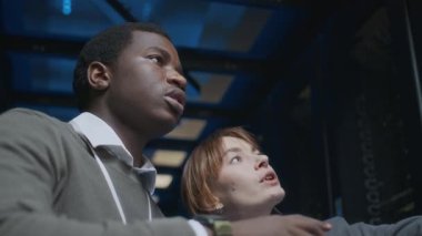 Genç, özgüvenli Afrikalı Amerikalı bilişim teknisyeninin, genç beyaz kadın meslektaşıyla bilgisayar kullanarak, veri merkezinde birlikte çalışırken görüntüsü düşük açılı.