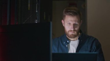 PC ve dizüstü bilgisayarda program kodlaması üzerinde çalışan beyaz erkek bilişim uzmanı yavaş yavaş portresi, sonra hafif bir gülümsemeyle kameraya bakıyor, veri merkezinde sunucu odasında oturuyor.