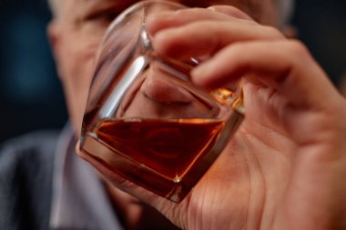 Zarif bir viski bardağını tutan ve karmaşık detaylar sunan ellerin yakın çekimi. Zengin kehribar likörüne odaklanmış, dokuları ve yansımaları vurguluyor.