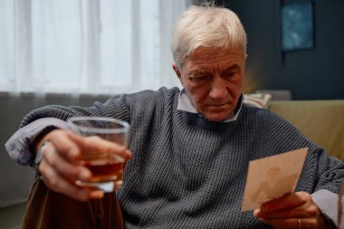 Kanepede oturan, elinde içki tutan ve eski fotoğraflara bakan düşünceli bir yüz ifadesi olan, süveter giyen, arka planda pencereden süzülen güneş ışığı olan yaşlı bir adam.