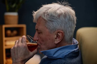 Yaşlı adam rahat bir ortamda, rahat bir şekilde koltukta dinlenirken içmekten zevk alıyor, sakin ve içerik tavrı sergiliyor.