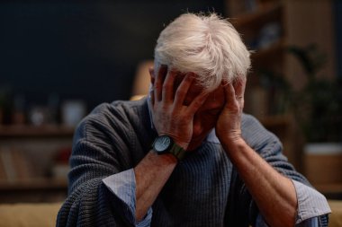 Yaşlı adam evde otururken yüzünü kapatıyor, üzüntü ve stresi ifade ediyor, arka planda kitaplık ve dekorla kaplı rahat bir odada saat ve süveter giyiyor.