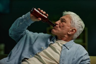 Yaşlı bir adam, rahatlama ifadesiyle koltukta otururken bira içiyor. Kısa beyaz saçlı kot gömlek giyiyor, anın tadını çıkarıyor.