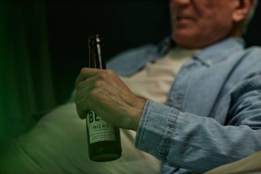 Otururken elinde bira şişesi tutan orta yaşlı bir adam, sıradan bir kot gömlek giyiyor. El ve şişeye odaklan, yüzünü odaklama, rahatlamış atmosferi taşıma.