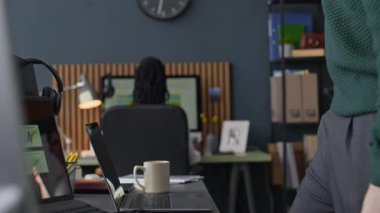 Günlük giysili ve gözlüklü genç beyaz bir adam ofis masasında oturuyor ve iş başında arka planda bilgisayar üzerinde çalışan meslektaşıyla birlikte dizüstü bilgisayar kullanıyor.