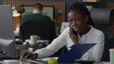 Beyaz tişörtlü Afro-Amerikan bir kadının cep telefonunun önünde otururken ve sözleşmeyi görüşmeyi bitirirken, meslektaşıyla açık alanda çalışırken orta ölçekli resmi.