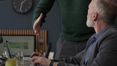 Gözlüklü ve günlük giysili genç beyaz adam ofis masasında olgun beyaz bir adama katılıyor ve iş projelerini bilgisayarda tartışmaya başlıyor, açık alanda birlikte çalışıyorlar.