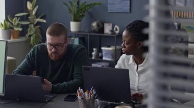 Orta boy beyaz adamın akıllı telefonu ofis masasında dizüstü bilgisayarların önünde genç siyahi bir kadın meslektaşıyla birlikte çalışırken görüntüsü.