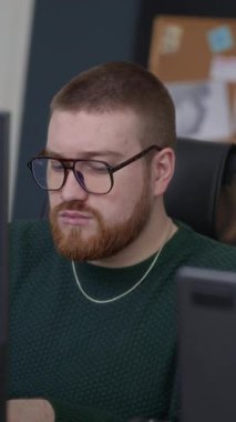 Dikey yavaş çekimde, gözlüklü, gözlüklü, genç, beyaz erkek çalışanın bilgisayar önünde masasında otururken, gündüz vakti ofiste çalışırken.