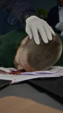 Steril eldiven konusunda tanınmayan bir adli tıp uzmanının dikey görüntüsü. Ofis masasında tanınmayan bir meslektaşla birlikte inceleme sürecini başlatmak için eldiven takan genç bir erkek kurbanı inceliyor.