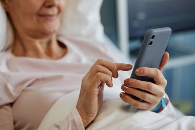 Akıllı telefon kullanırken gülümseyen, rahat kıyafetler giyen ve rahatlatan yaşlı bir kadın. Elindeki cihazla mutlu ve memnun görünüyor.