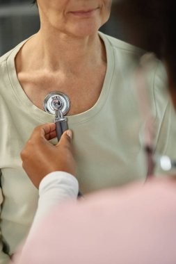 Doktor steteskop kullanarak hastayı muayene ediyor, kalp atışlarını ve akciğer seslerini kontrol ediyor. İşlem sırasında hastanın sakin bir tavır sergilediği sağlık hizmetlerine odaklanın