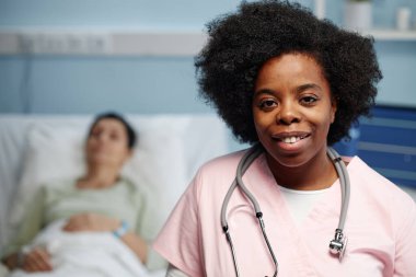 Portrait of female African American doctor smiling confidently with stethoscope, patient lying in bed in background, hospital setting showing care and professionalism clipart