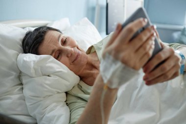 Yaşlı bir kadın hastane yatağında uzanıyor, gülümsüyor ve kolunda serumla akıllı telefona bakıyor, neşe ve bağlantı anı yaratıyor.