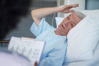 Yaşlı adam hastane yatağında rahat bir şekilde dinleniyor, elini kaldırıyor ve sıcak bir şekilde gülümsüyor. Hemşire ön planda pano tutuyor.