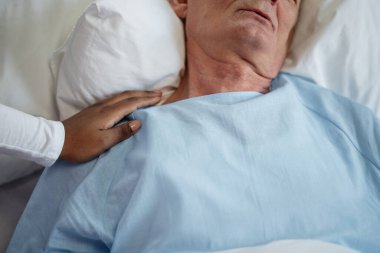 Hasta yatağında yatan yaşlı hastayı rahatlatan Afro-Amerikan kişi sakin görünüyor, omuz omuza destek oluyor, hastane ayarları görülebilir.