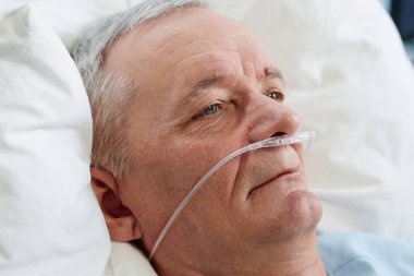Yaşlı adam hastane yatağında dinleniyor, burun kanülünden oksijen alıyor, düşünceli ve sakin görünüyor, arka plan yumuşak yastıklarla dolu, sakin bir atmosfer yaratıyor.
