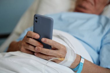 Hastane yatağında yaslanan yaşlı kişi, yatarken akıllı telefonu tutuyor, kolunda tıbbi bileklik ve damar yolu ile iyileşme veya dinlenme safhasını gösteriyor.
