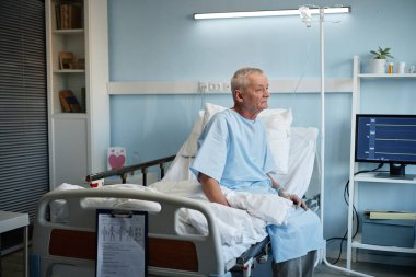 Yaşlı bir erkek hasta hastane yatağında, çevresinde tıbbi malzemelerle aydınlık bir odada tedavi görüyor.