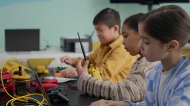 Modern sınıftaki masada oturan çeşitli ilkokul öğrencilerinin dizüstü bilgisayarlarıyla robotlar yaptığı orta boy bir fotoğraf.