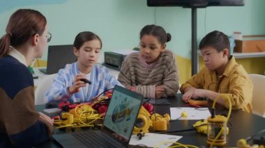 Genç, beyaz kadın öğretmenin gündelik kıyafetleriyle, Alfa Genelkurmay 'ındaki çoklu etnik çocuklarla aynı masada otururken ve bilgisayar ekranında dijital çizim kullanarak robot nasıl yapılır açıklarken orta boy fotoğrafı.