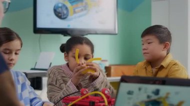 Çok ırklı Gen Alpha öğrencilerinin, STEM dersi sırasında öğretmenleriyle masaya oturup yeni beceriler kazandıkları orta boy bir fotoğraf.