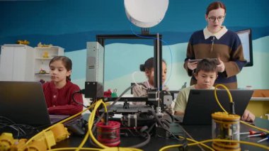 Modern iyi donanımlı sınıflarda dizüstü bilgisayarların önünde oturan çok ırklı çocukları eğiten, üç boyutlu baskı ve programlamayı denetleyen Kafkas kadın öğretmeninin bıçaklı fotoğrafı.