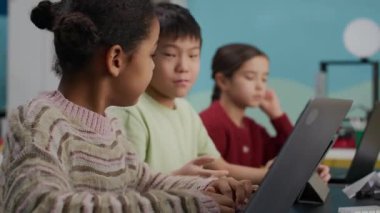 Yan görüş, sınıftaki melez bir kızın, Asyalı bir oğlanla, dizüstü bilgisayarların önünde oturup modern, iyi donanımlı sınıflarda programlama çalışırken görüntüsü.