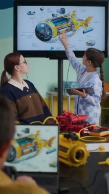 Dijital tablet taşıyan zeki Asyalı bir kızın modern iyi donanımlı sınıflar konusunda robotik dersinde duvardaki şemayı kullanarak genç beyaz kadın öğretmene robot yapısı hakkında rapor vermesi dikey.