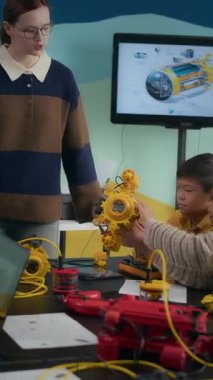 Genç, beyaz kadın öğretmenin, robot sınıfı sırasında masada oturan meraklı Alfa Asyalı erkek ve melez kıza sarı robot modeli göstermesinin dikey görüntüsü.