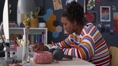 Yan görüntüde, parlak kazak giyen Afro-Amerikalı genç bir kızın okul ödevini yaparken rahat odasında otururken fotoğraf çekimi var.