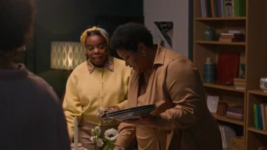 Genç nesil aile üyeleriyle sohbet ederken çiçeklerle ve mumlarla süslenmiş yemek masasında bulaşık takımı servis eden orta boy siyahi bir kadın.