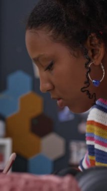 Okuma ödevini tamamlayan, arka planda bir sürü kitapla birlikte odasında oturan Afro-Amerikan ergen kızın dikey ve yavaş çekimi.