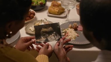 Bebeğin ultrason görüntüsüne bakıp fetüs gelişimini gösteren eşlerin omuz üstünden çekimleri evde kutlama sırasında şenlikli yemek masasında otururken.