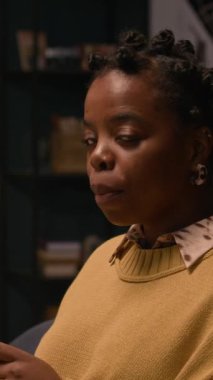 Siyahi kadının mesajlaşmasının dikey portresi. Sonra hafif bir gülümsemeyle kameraya dönüyor. Oturma odasında yumuşak, ılık bir ışıkla çekiliyor.