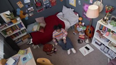 Yüksek açılı, üzgün, Afrika kökenli Amerikalı bir kızın üst düzey bir iç mekan ile odasına girmesi, yerde oturması ve kendini yalnız hissetmesi, dizlerine sarılması ve güvensiz hissetmesi.