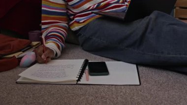 El kamerasıyla, parlak günlük giysili, ev ödevi yapan, okul ödevi yazan ve odasında ders çalışmak için dizüstü bilgisayar kullanan siyahi bir kızın el kamerası görüntüsü.