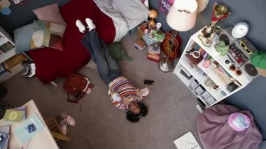 Siyahi genç kızın rengarenk kıyafetleri içinde ağır çekimde yerde yatıp kulaklıkla müzik eşliğinde dans etmesi odasında maksimum içsellikle dinlenmesi evde tek başına vakit geçirmesi.