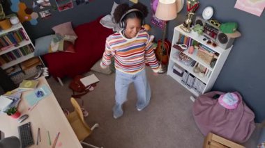 Yüksek açılı yavaş çekim mutlu siyah genç kız rengarenk günlük giyimli kulaklıkla müzik eşliğinde dans ediyor, odasında maksimum iç mekan ve duvarlarda bir sürü posterle yalnız vakit geçiriyor.