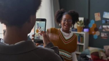 Siyah gencin akıllı telefondaki dans eden kız arkadaşının videosunu çekerken yavaş çekimleri sosyal medya için evde rahat bir odada moda içeriği yaratıyor.