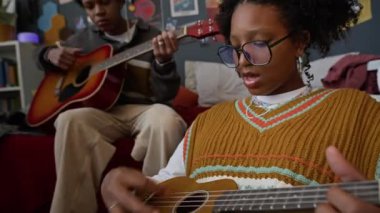 Yetenekli Afro-Amerikan gen z kızı, gözlüklü, erkek arkadaşıyla gitar çalıp şarkı söylerken ve gitar çalarken el kamerasıyla çekilmiş.