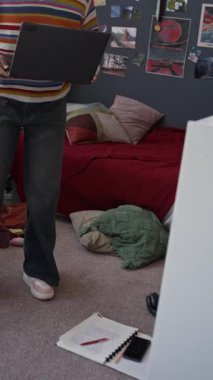 Dikey olarak, parlak günlük giysili genç bir kızın duvarlarında bir sürü posterle odasına girişinin dizüstü bilgisayarla oturuşunun yataktan yatağa oturuşunun ve daktiloyla yazışmasının fotoğrafı.