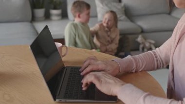 İki çocuğu arka planda çalarken beyaz bir kadının masada oturup dizüstü bilgisayarına klavye yazarken orta boy fotoğrafı.